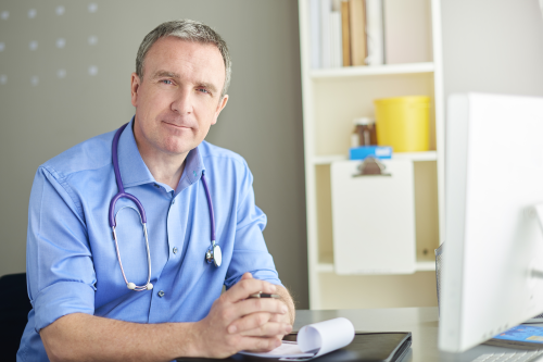 A male doctor sits at a desk in front of a computer. He is wearing a stethoscope and has his fingers laced in front of him.