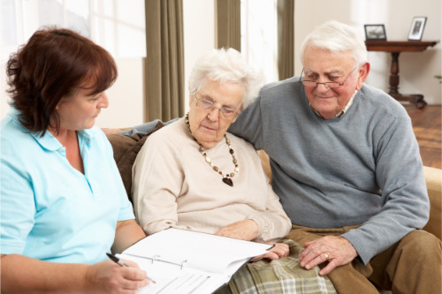 An elderly couple sit on a sofa, with a lady holding a ringbinder file. She is holding a pen and talking to the couple.