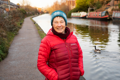 A woman is walking down a canal path. She is smiling at the camera, and is wearing a woolly hat and a red puffer coat.