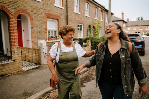 Two women walk down a residential street. They are laughing. The woman on the left is wearing khaki green dungarees, and the woman on the right is wearing all black besides a denim jacket.