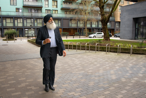 An older man is walking outside past an office building. He has a long white beard, and is wearing a suit and a turban.