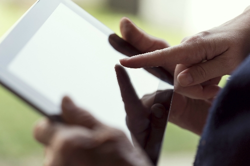 A pair of hands hold a tablet device, whilst another hand points at the screen.