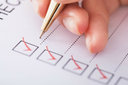 A close-up photo of a hand holding a pen, resting on a piece of paper with 4 boxes printed on. Each box has a red tick mark inside.