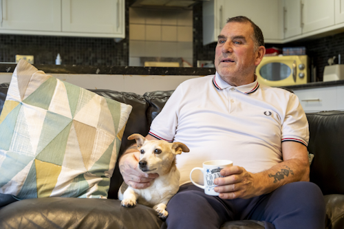 A man wearing a polo shirt and jeans sits on his sofa, with a small terrier dog next to him. He is stroking the dog, and holding a mug of tea in his other hand.