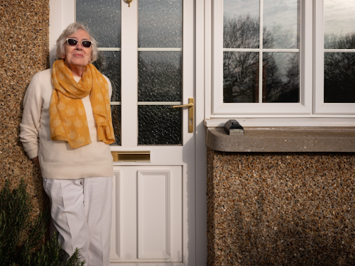 An elderly woman is standing outside by the door to her house. She is wearing white trousers, a cream jumper, a golden yellow scarf, and dark sunglasses.