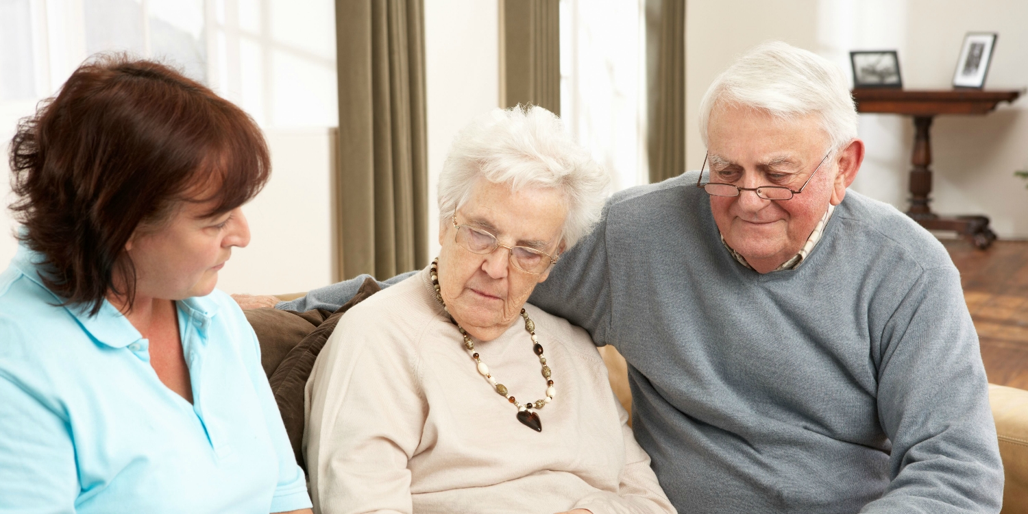 An elderly couple sit on a sofa, with a lady holding a ringbinder file. She is holding a pen and talking to the couple.