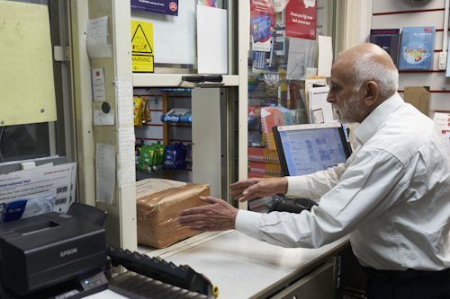 A man with grey hair wearing a white button-down shirt sits behind a counter at the Post Office. A parcel sits on the desk, and he is reaching out to take it.