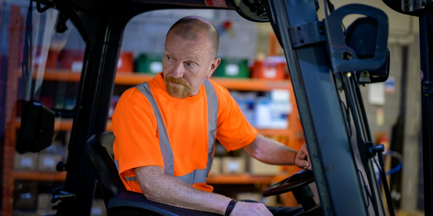 A man operates a forklift, he is wearing a hi-vis t-shirt